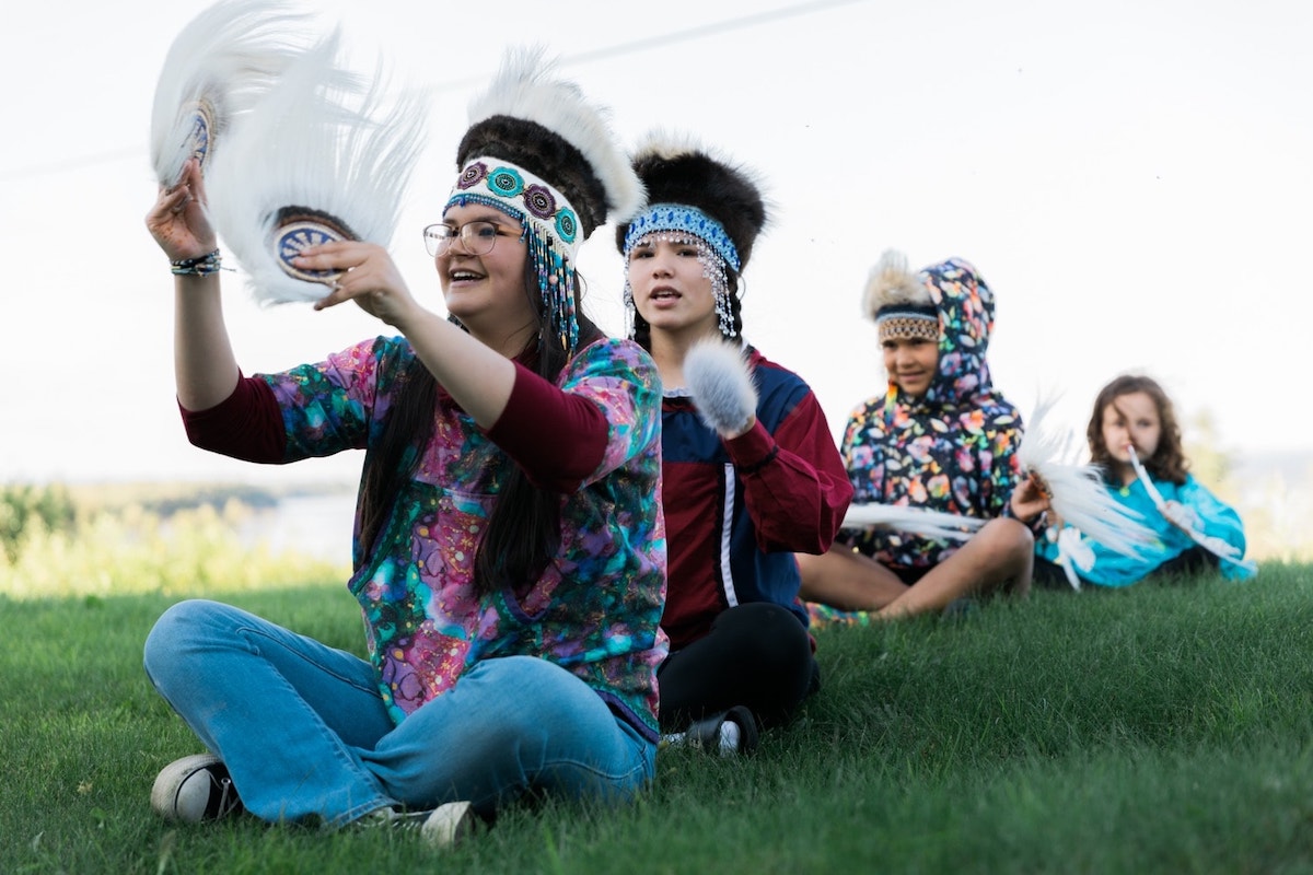 oung dancers in vibrant traditional regalia with beaded headdresses and fur perform a cultural dance on a lush green lawn, their feathered fans raised in a moment of joyous expression.